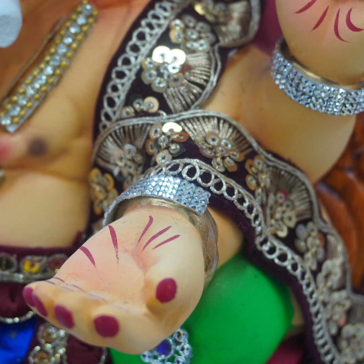 Close-up of a decorative Ganesha idol showing the face, decorated arms, and ornate silver and gold jewelry against a colorful background.