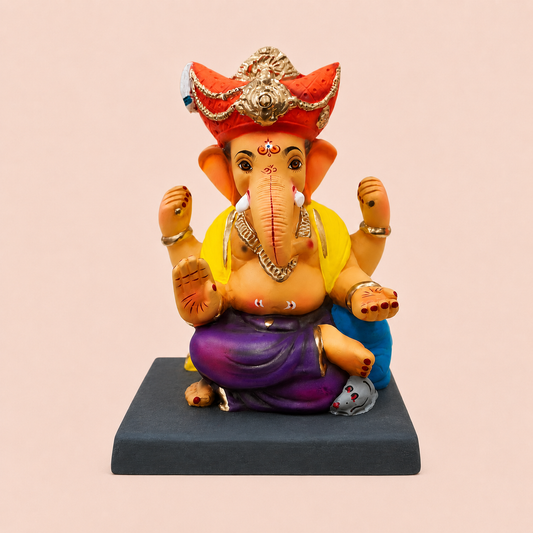 A painted Ganesha statue seated on a dark rectangular base against a pale pink background. The statue has a peach-toned body, a red and gold ornate crown, a yellow upper garment, a purple lower garment, and blue pants.